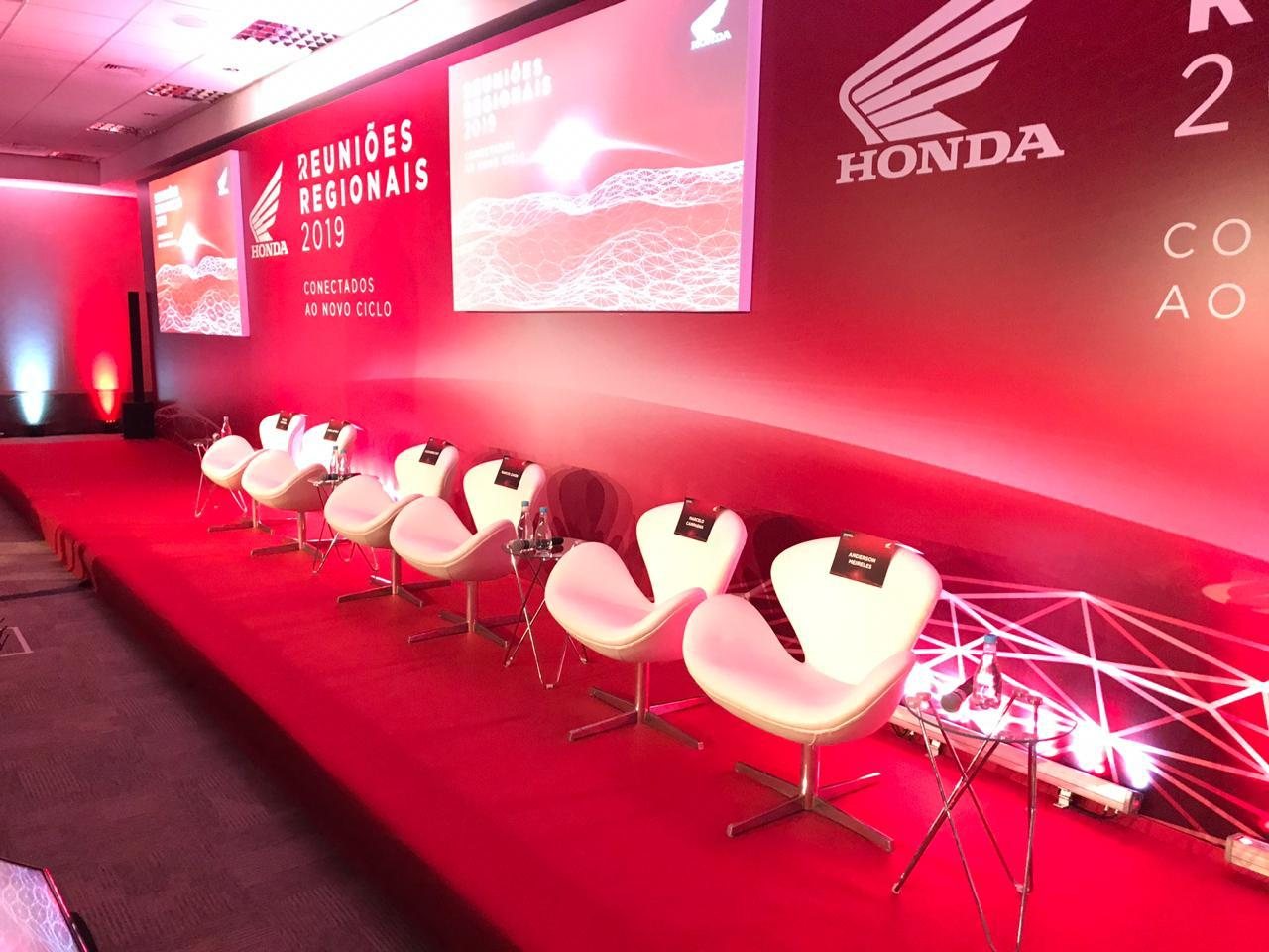 Plenária Honda Reuniões Regionais com palco iluminado em vermelho, poltronas brancas alinhadas e telas de projeção ao fundo