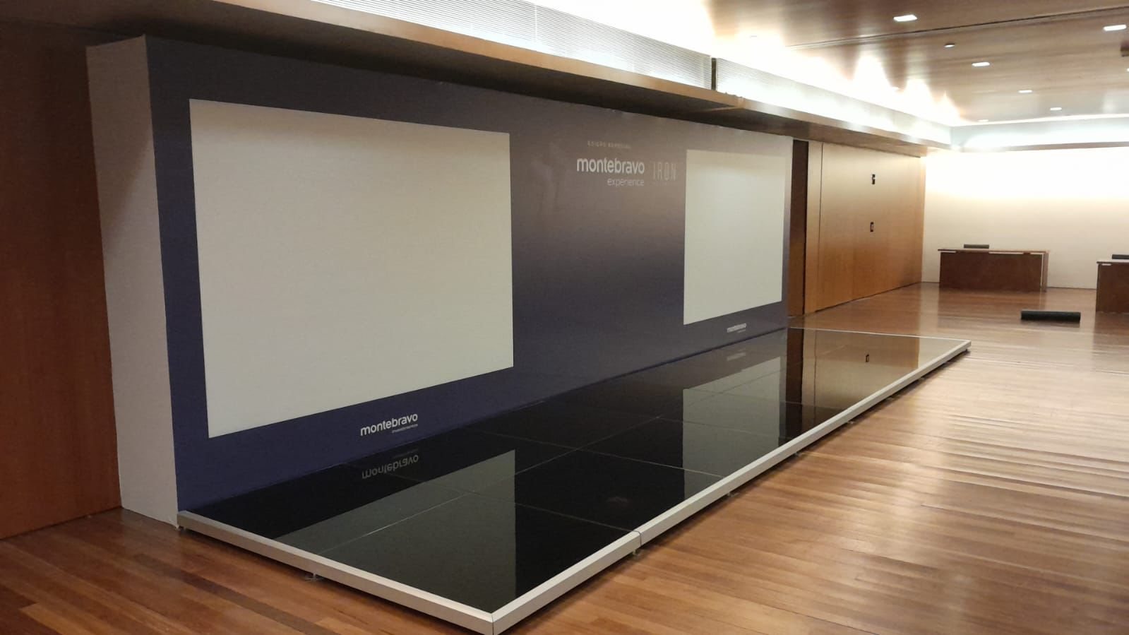 Palco de vidro preto espelhado do Montebravo Experience com fundo em painel azul-escuro e duas áreas de projeção
