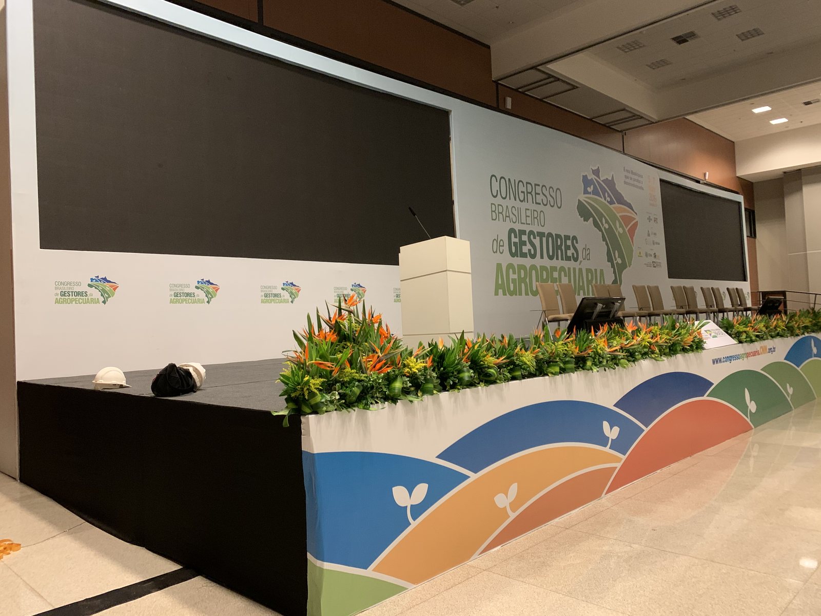 Palco do Congresso Brasileiro de Gestores da Agropecuária com mesa diretora, paisagismo de flores tropicais e painel impresso